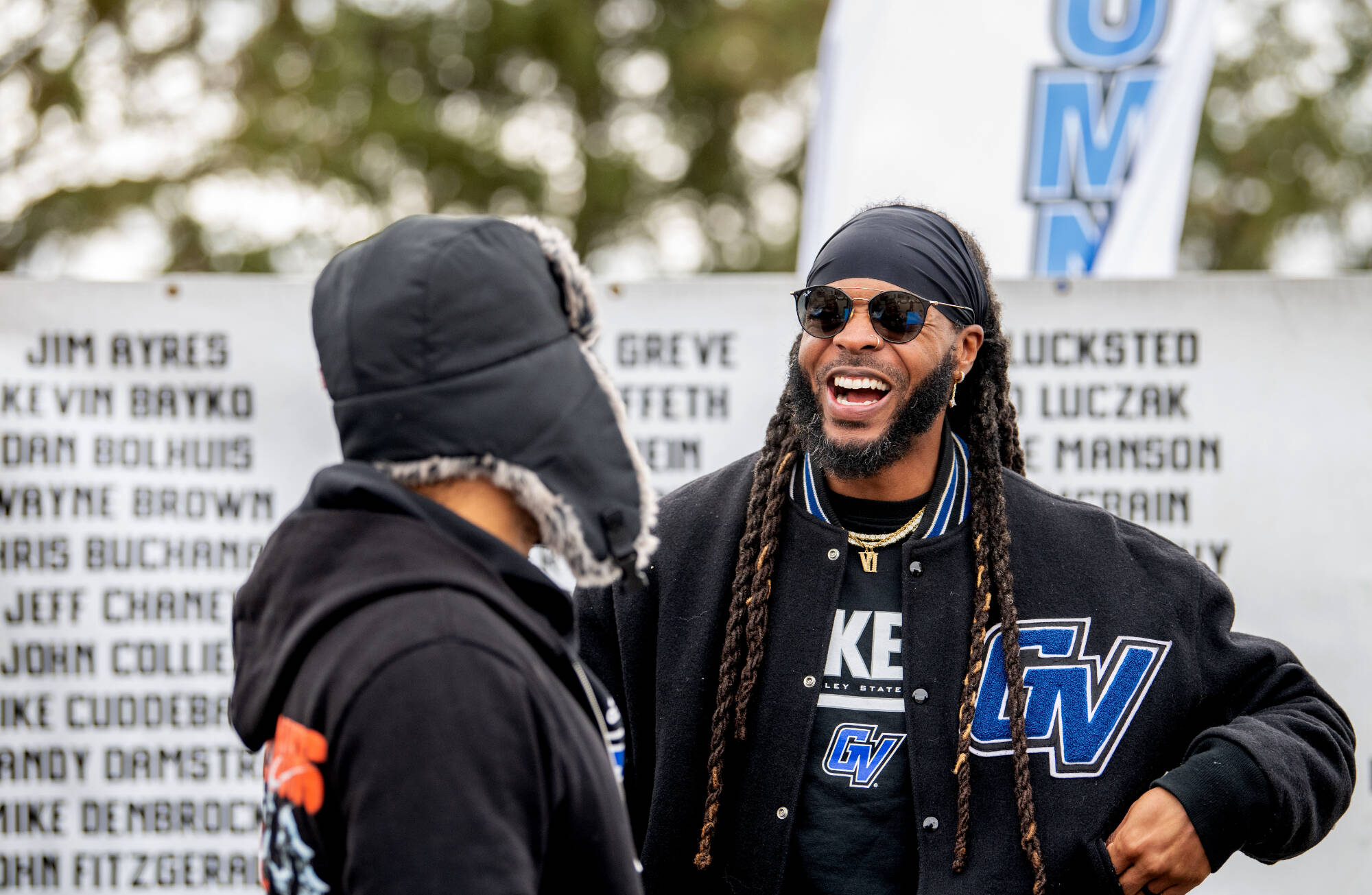 Two students laughing and talking outdoors at a homecoming event, wearing black varsity jackets with GV letters. Behind them is a white memorial wall with names engraved on it and a banner with blue letters visible in the background. One student wears a b